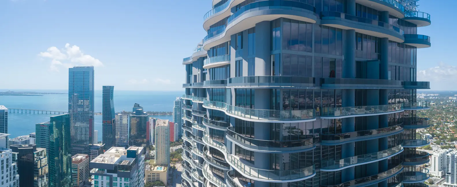 Brickell Flatiron's curved glass facade overlooking Miami skyline and waterfront.